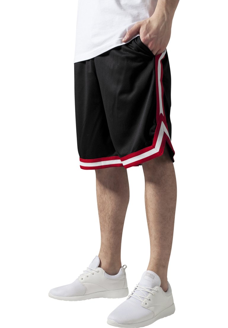 Personne portant un short de sport noir avec des bandes rouges et blanches et des baskets blanches, debout avec une main dans la poche.