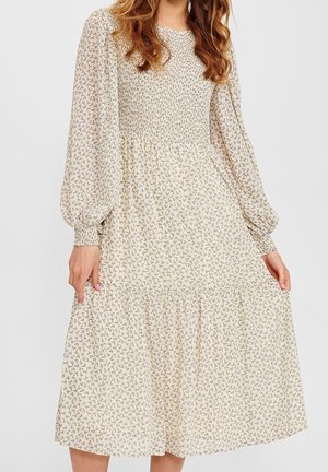 Kobieta ubrana w kremową sukienkę midi z długim rękawem, z marszczonym gorsetem i małym fioletowym kwiatowym wzorem, delikatnie trzymająca boki spódnicy.