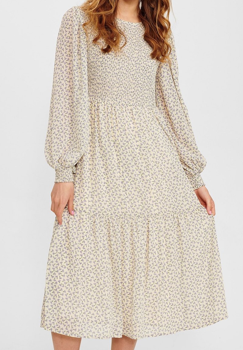 Femme portant une robe midi crème à manches longues avec un corsage froncé et un petit motif floral violet, tenant doucement les côtés de la jupe.