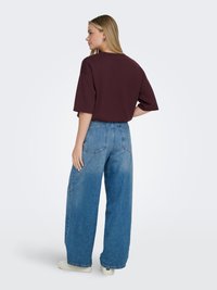 Femme aux cheveux blonds portant un haut ample à manches courtes bordeaux, un jean à jambes larges bleu et des baskets blanches, debout devant un fond uni.