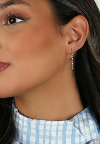 Guldtonade hängande örhängen med små klara ädelstenar, i en elegant kedjedesign. Örat visas framträdande.