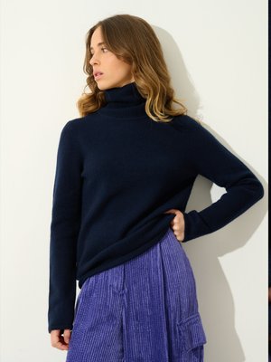 Maglione a collo alto di colore blu navy, realizzato in un tessuto morbido, con maniche lunghe. Abbinato a una gonna plissettata in corduroy viola con tasche.