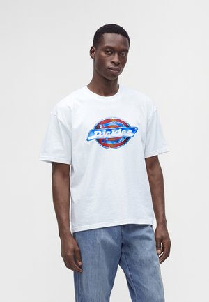 Мъж, стоящ, облечен в бяла тениска с логото на Dickies и светлосини дънки пред светъл едноцветен фон.