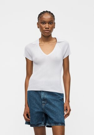 Frau trägt ein weißes, kurzärmliges, geripptes V-Ausschnitt-Top mit Kragen und hoch taillierte blaue Denim-Shorts vor einfachem Hintergrund.