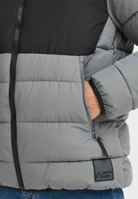 Šedá a čierna puffa bunda s prešívanou textúrou, so zapínaním na zips vpredu, bočnými vreckami a čiernym logom pri leme.