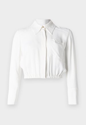 Blouse blanche courte avec des manches longues, un col pointu, une fermeture à boutons à l'avant et une seule poche sur la poitrine. Tissu lisse et léger.