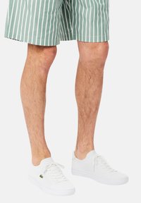 Białe siateczkowe trampki z płaskimi sznurowadłami i zielonym haftowanym logo, zestawione z zielonymi spodenki w paski. Skoncentrowane na lekkiej, oddychającej konstrukcji.