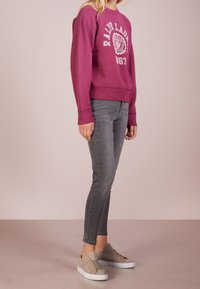 Lila sweatshirt med vit logotyp och årtal på framsidan, kombinerad med grå smala jeans och ljusbeige sneakers med texturerad design.