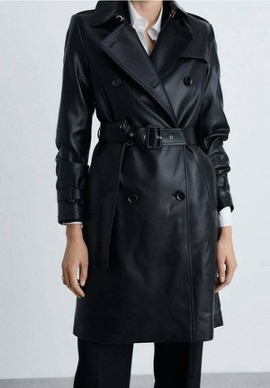 Kobieta nosząca czarny, dwurzędowy płaszcz trencz z eko skóry z paskiem, na białej koszuli i czarnych spodniach na jednolitym tle.