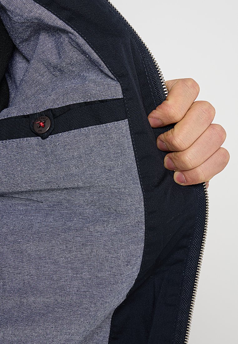 Intérieur d'une veste navy avec un tissu texturé bleu et gris, un détail de bouton avec un accent rouge, et une ouverture zippée.