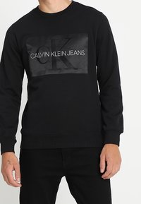 Man i svart Calvin Klein Jeans sweatshirt med stort "CK"-logo och vit text på bröstet, tillsammans med svarta byxor.