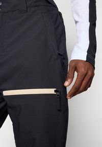 Fekete sínadrág texturált anyagból, kontrasztos bézs cipzáras zsebbel, funkcionális derékkal és sima varrási részletekkel.