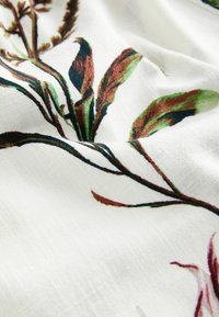 Tissu blanc avec un motif botanique présentant des feuilles brunes et vertes ainsi que de fines branches, montré en gros plan avec une texture et des plis visibles.