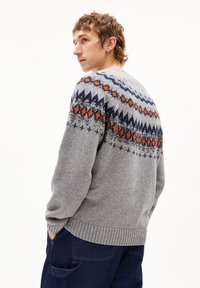 Grauer gestrickter Pullover mit dekorativen blauen, orangefarbenen und marineblauen Mustern über den Schultern. Rippstrickbündchen und Saum. Über blauen Hosen getragen.