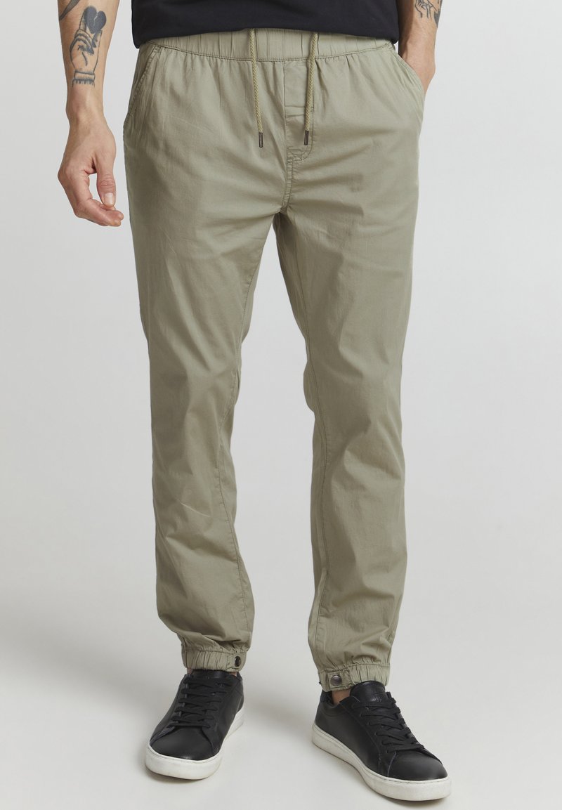 Pantalons jogger beige clair avec une taille élastique et des poignets élastiques, dotés de poches latérales et de détails en cordon de serrage. Associés à des baskets noires.