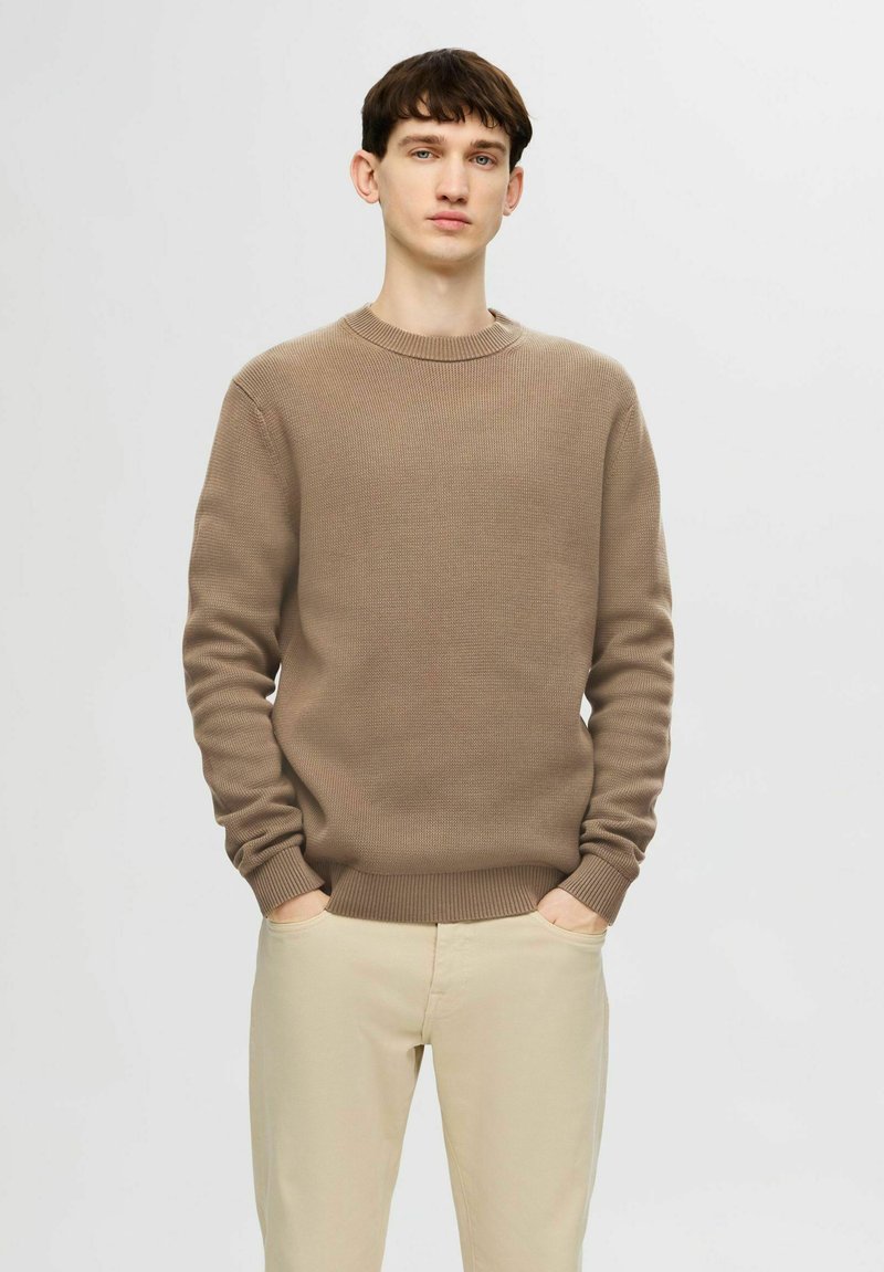 Maglioncino beige lavorato a maglia con collo a girocoscia e polsini a costine. Il modello sta in piedi con le mani in tasca, indossando pantaloni beige chiaro.