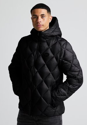 Jeune homme portant une veste matelassée noire à capuche, les mains dans les poches, debout devant un fond gris clair uni.