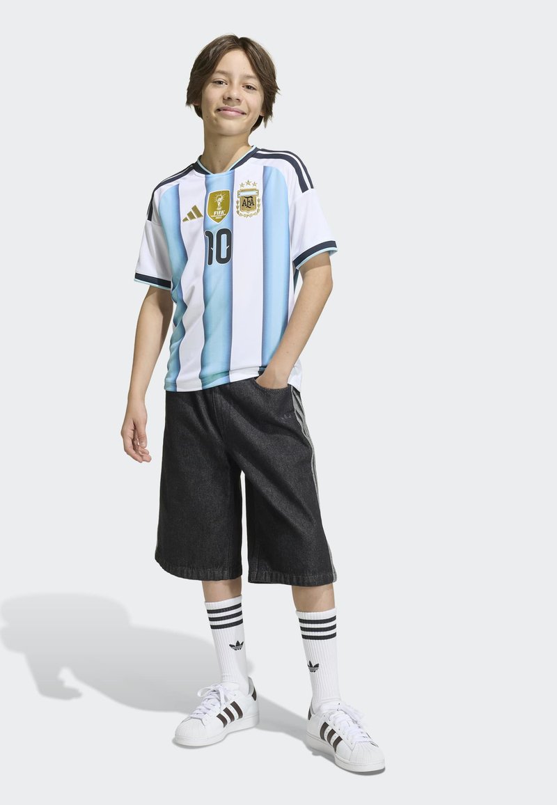 Jeune personne se tenant avec assurance, portant un maillot de soccer de l'Argentine, un short noir, des chaussettes blanches Adidas et des baskets, sur un fond uni.