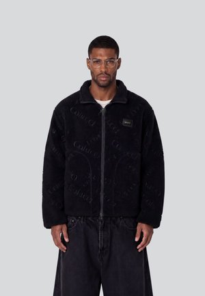 Mann mit klaren Brillengläsern, einer schwarzen Fleecejacke mit Reißverschluss und dezentem Textmuster sowie lockeren schwarzen Hosen, stehend vor einem einfachen hellen Hintergrund.
