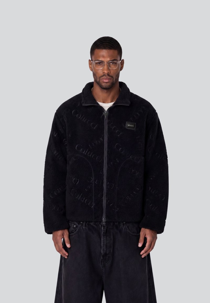 Homme portant des lunettes transparentes, une veste polaire noire zippée avec un motif de texte subtil et un pantalon noir ample, debout sur un fond clair uni.