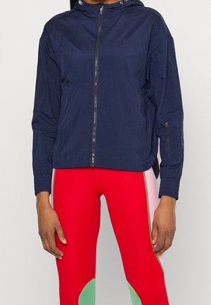 Persona con chaqueta con capucha y cremallera azul marino y leggings rojos con paneles de bloque de color rosa y verde, de pie frente a un fondo liso.