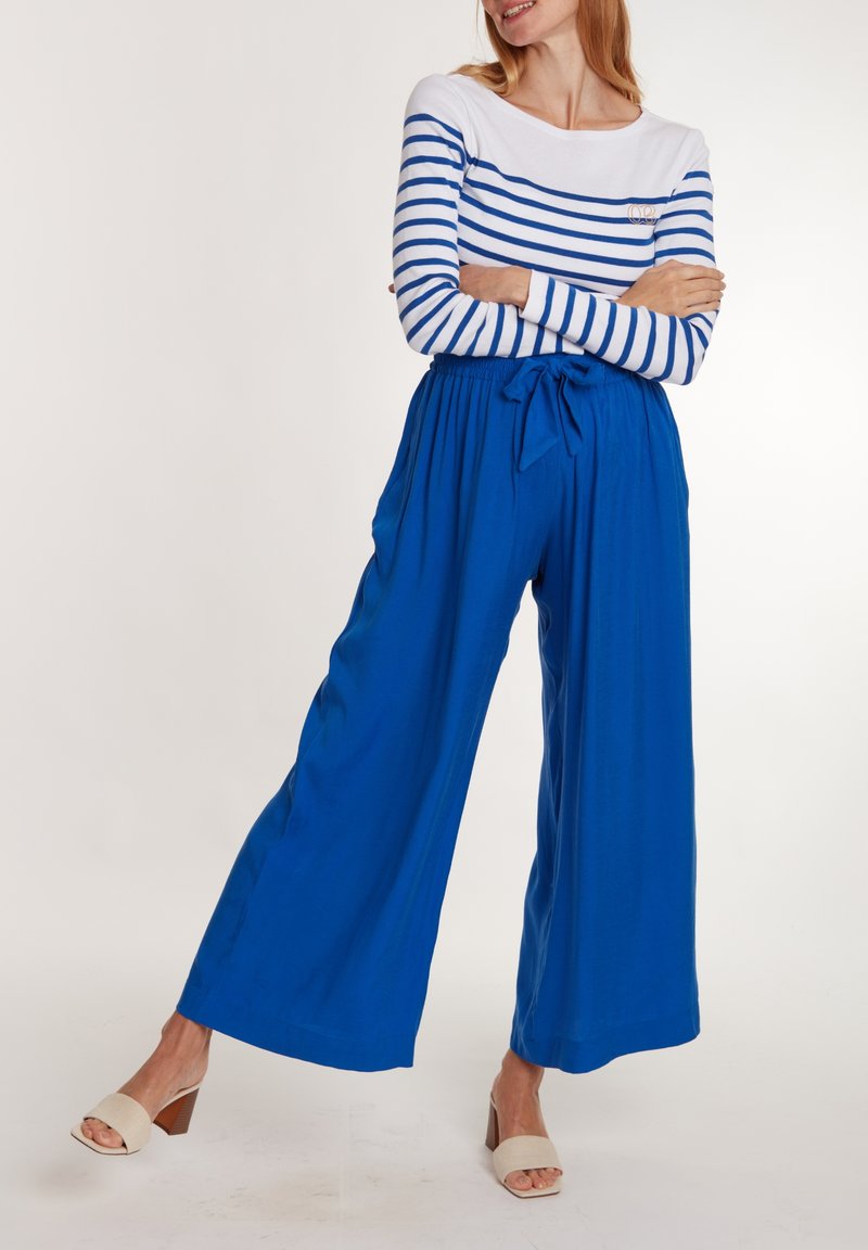 Pantalons larges bleus avec une ceinture nouée, associés à un haut à rayures blanc et bleu à manches longues, et des sandales à talons beiges.