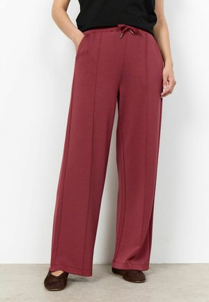 Vrouw die hoge taille wijde bordeauxrode broek draagt met zijzakken en trekkoord, gecombineerd met een zwarte top en donkerbruine schoenen.