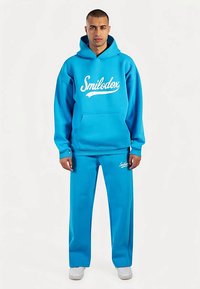 Conjunto deportivo de dos piezas en azul brillante. Incluye una sudadera con capucha con un logo blanco y pantalones deportivos a juego con bolsillos laterales.
