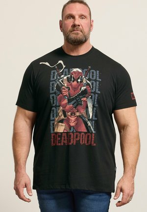 Homme barbu portant un t-shirt noir Deadpool avec Deadpool croisant les bras tenant des épées, debout devant un fond uni.