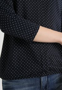 Černá blůza s puntíky bílé barvy, tříčtvrteční rukávy, zaoblený spodní lem a měkká textilie, stylizovaná s modrými džíny.