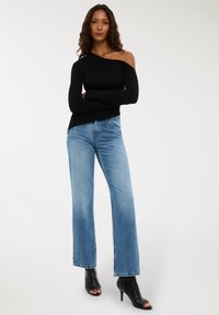 Kobieta z długimi kręconymi włosami, ubrana w czarny sweter z odkrytymi ramionami, jasnoniebieskie jeansy o prostym kroju i czarne odkryte wysokie obcasy.