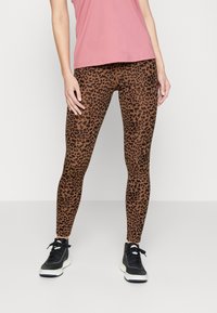 Leggings a stampa leopardata in marrone con macchie nere, design aderente e tessuto elastico, abbinati a scarpe sportive nere e a una maglietta rosa.