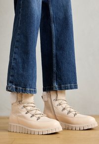Botas de tobillo beige con suela de goma, que presentan cordones, ojales de metal y acentos esponjosos en el cuello. Combinadas con jeans de denim anchos.