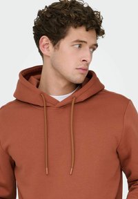 Jonge man met krullend haar, gekleed in een roestkleurige hoodie over een wit shirt, kijkt naar rechts tegen een effen achtergrond.