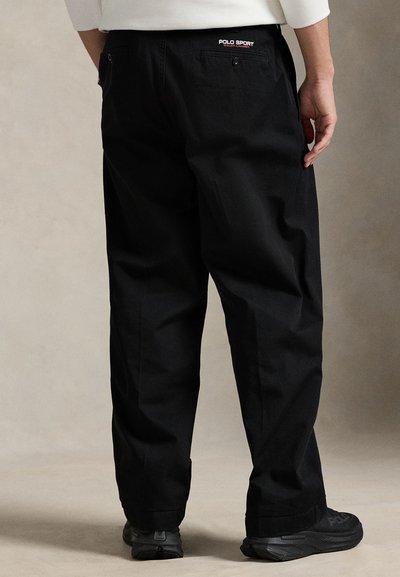 Μαύρο βαμβακερό παντελόνι με άνετη εφαρμογή, με λογότυπο στο πίσω μέρος, πλαϊνές τσέπες και ομαλή υφή. Φορεμένο με μαύρα παπούτσια.