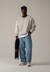 Szürke pulóver "BLKVIS NY" felirattal, kék farmernadrág, alatta fehér ing, tengerészkék sapka, kezében tengerészkék sál narancssárga részletekkel. Fehér tornacipő.