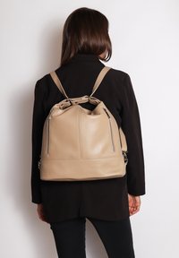 Zaino a secchiello in pelle beige con due cerniere laterali, chiusura superiore a cordoncino e spallacci regolabili. Texture liscia e forma strutturata.