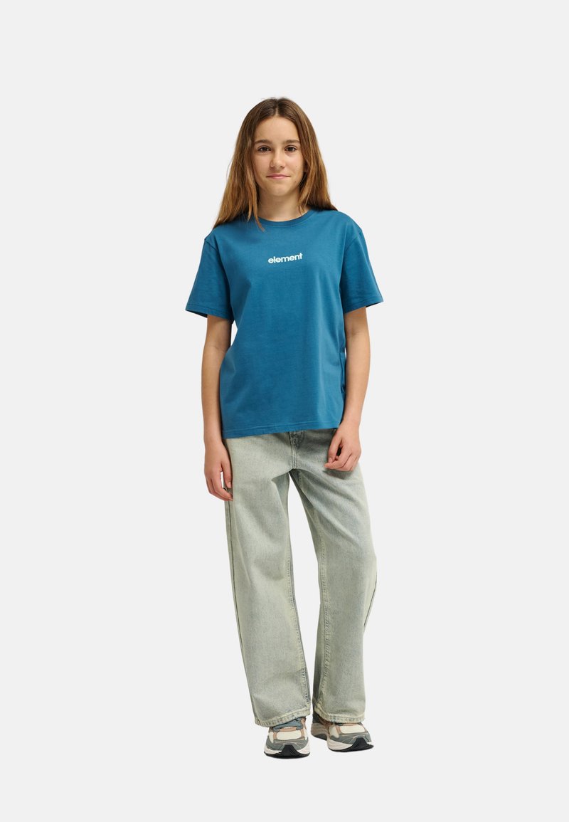 Joven de pie con camiseta informal color verde azulado con el logo "element", pantalones vaqueros anchos de pierna clara y zapatillas deportivas, frente a un fondo blanco liso.