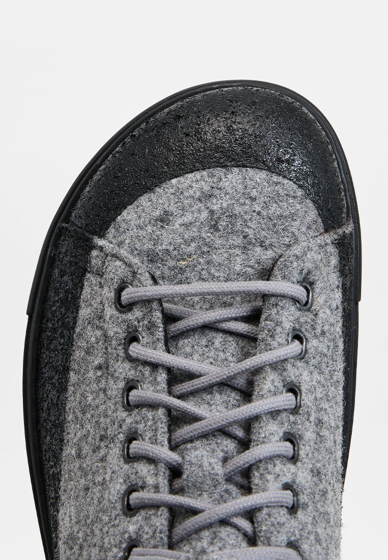 Zapatilla gris de felpa con puntera de goma negra, cordones redondos y superficie texturada. Presenta detalles cosidos y ojales reforzados.