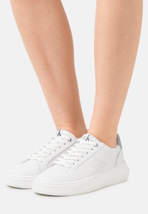 Weiße Sneakers mit niedrigen Schaft und weißen Schnürsenkeln, das "cK"-Logo an der Seite und Zunge, barfuß vor einem grauen Hintergrund getragen.