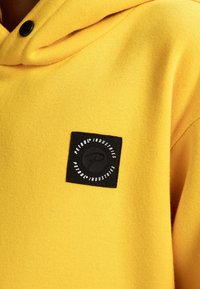 Felpa gialla con un patch quadrato nero sul petto che mostra il testo "Petrol Industries" all'interno di un cerchio bianco attorno a un logo nero.