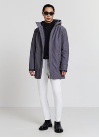 Cappotto grigio con cappuccio e cerniera frontale, due tasche laterali, indossato sopra un dolcevita nero, abbinato a pantaloni bianchi e stivaletti neri.