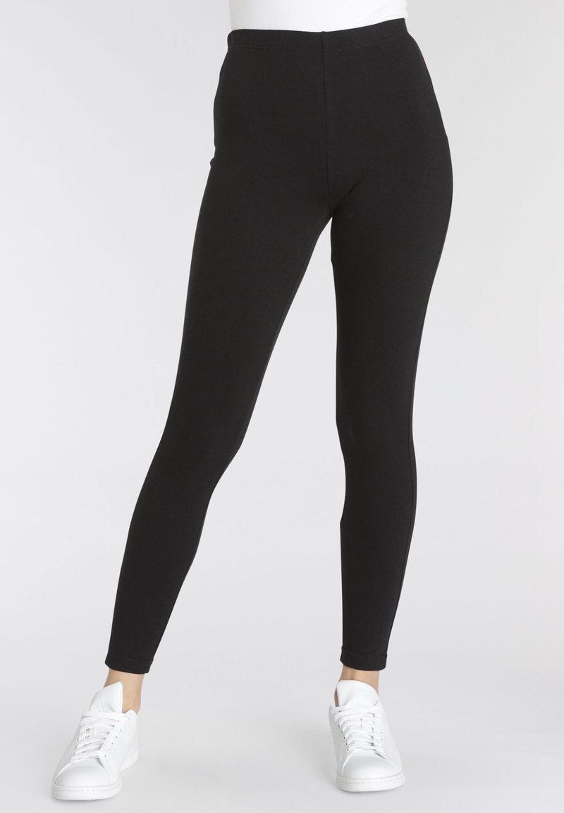 Leggings noir extensibles avec une taille haute, une coupe ajustée et une texture lisse, associés à des baskets blanches. Pas de logos ni de motifs visibles.