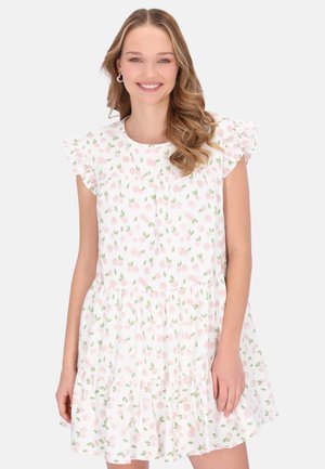 Smilende kvinde med bølget blondt hår iført en hvid kjole med lyserødt og grønt blomsterprint og flæseærmer, stående mod en hvid baggrund.