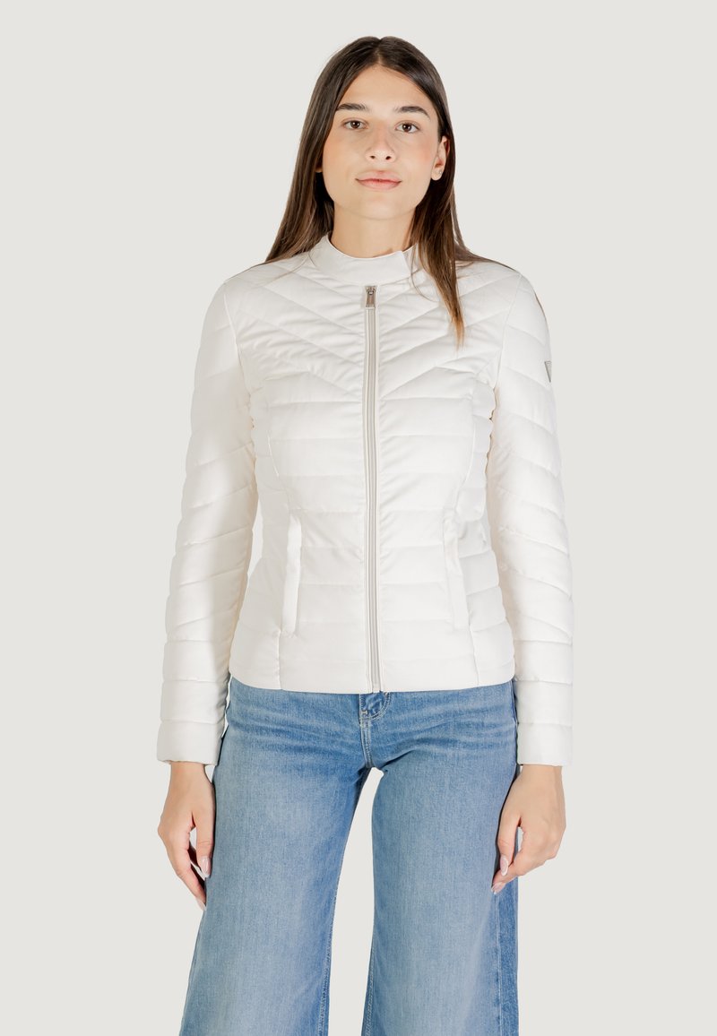 Giacca imbottita bianca trapuntata con colletto alto, zip frontale e design aderente. Indossata con jeans svasati blu. Texture liscia.