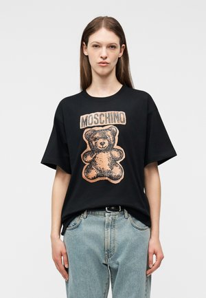 Vrouw met lang bruin haar die een zwart Moschino T-shirt draagt met een grote teddybeerprint en een lichtblauwe spijkerbroek.