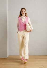 Vesta tricotată roz peste o bluză albă, purtată cu pantaloni bej și adidași roz. Ținând o geantă bej, pe un fundal gri deschis.