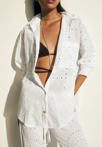 Chemise blanche boutonnée en tissu léger, avec des détails découpés. Associée à un haut de bikini marron. Texture subtile et coupe décontractée.