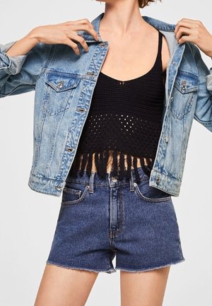 Frau trägt eine hellblaue Jeansjacke über einem schwarzen gehäkelten Crop-Top und hoch taillierten dunkelblauen Jeansshorts.