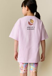 Rozā kokvilnas T-krekls ar īsām piedurknēm un aizmugurē grafiku ar ziedu un tekstu "AUZIET KĀ SAVVAĻAS ZIEDS". Kombinēts ar krāsainām legingām ar ziedu rakstu.